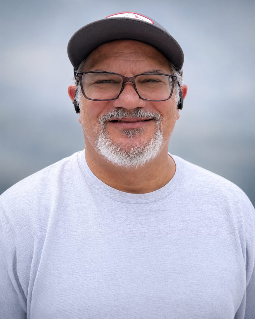 Casual outdoor portrait of a smiling man with glasses and a gray beard wearing a baseball cap, white sweatshirt, and wireless earbuds, posed against a softly blurred gray background.
