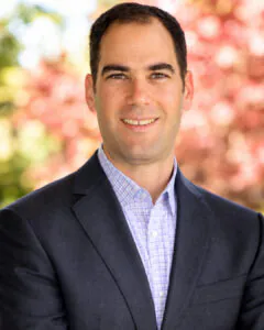 Professional outdoor headshot of a smiling man with short dark hair wearing a navy blazer and light purple checkered dress shirt, posed in front of a softly blurred background with green and pink foliage.