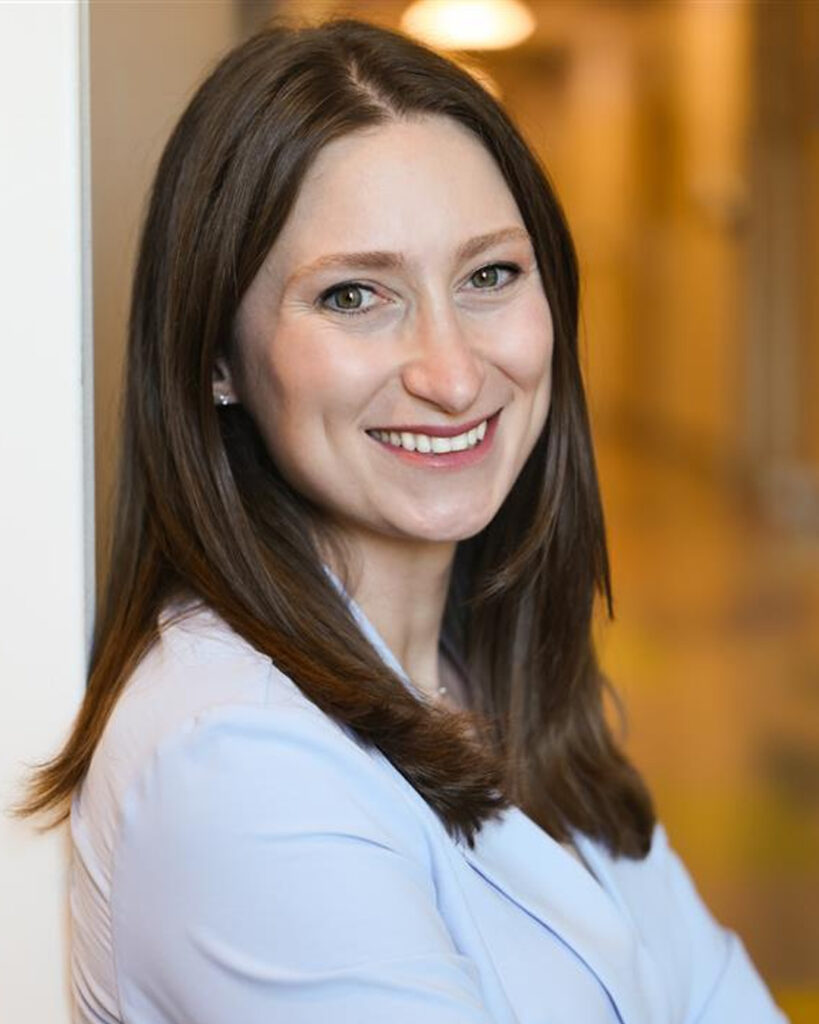 Portrait of a smiling woman with long brown hair, wearing a light blue blazer and standing with her arms crossed against a softly blurred indoor background.