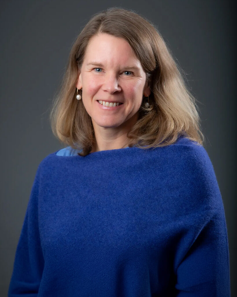 Headshot of a smiling woman with light brown hair, wearing a blue sweater, against a dark gray background.