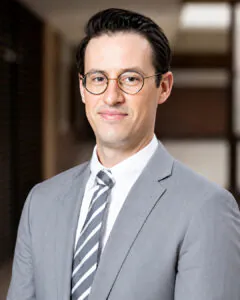 Professional headshot of a man wearing glasses, a gray suit, and striped tie, standing in an indoor setting.
