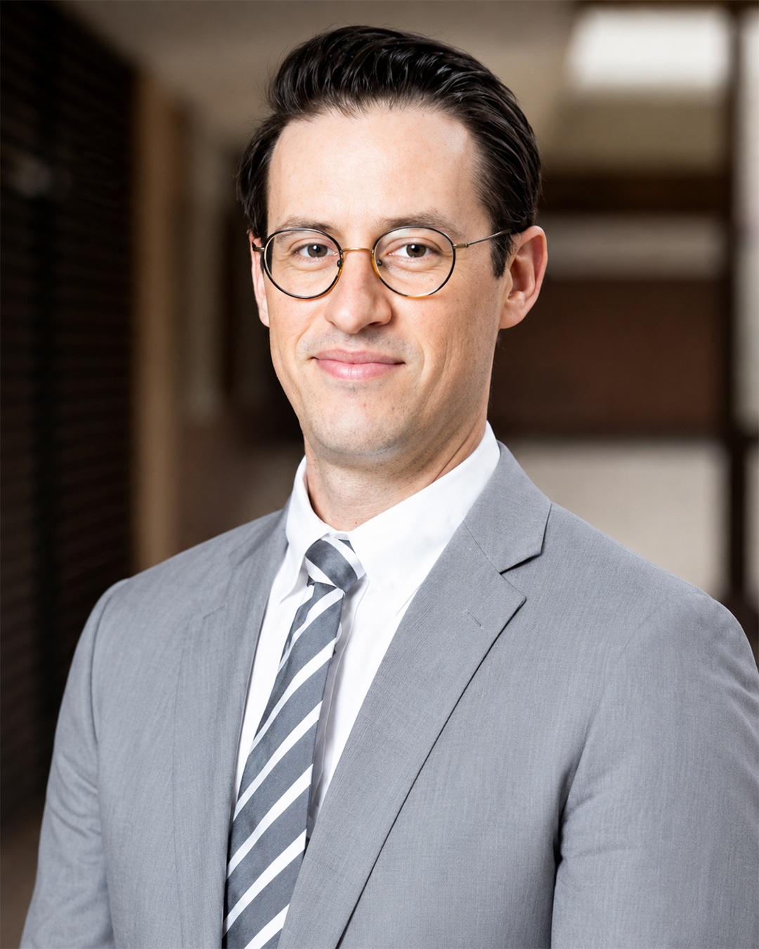 Professional headshot of a man wearing glasses, a gray suit, and striped tie, standing in an indoor setting.