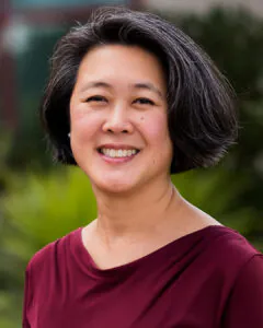 Headshot of a smiling woman with short dark hair, wearing a burgundy top, outdoors with greenery softly blurred in the background.