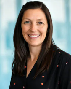 Headshot of a smiling woman with long dark brown hair, wearing a black blouse with a subtle pattern, against a softly blurred light blue background.