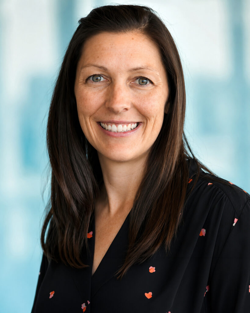 Headshot of a smiling woman with long dark brown hair, wearing a black blouse with a subtle pattern, against a softly blurred light blue background.