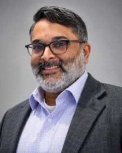 Headshot of a smiling man with short dark hair and a salt-and-pepper beard, wearing glasses, a light purple shirt, and a gray blazer, against a neutral background.
