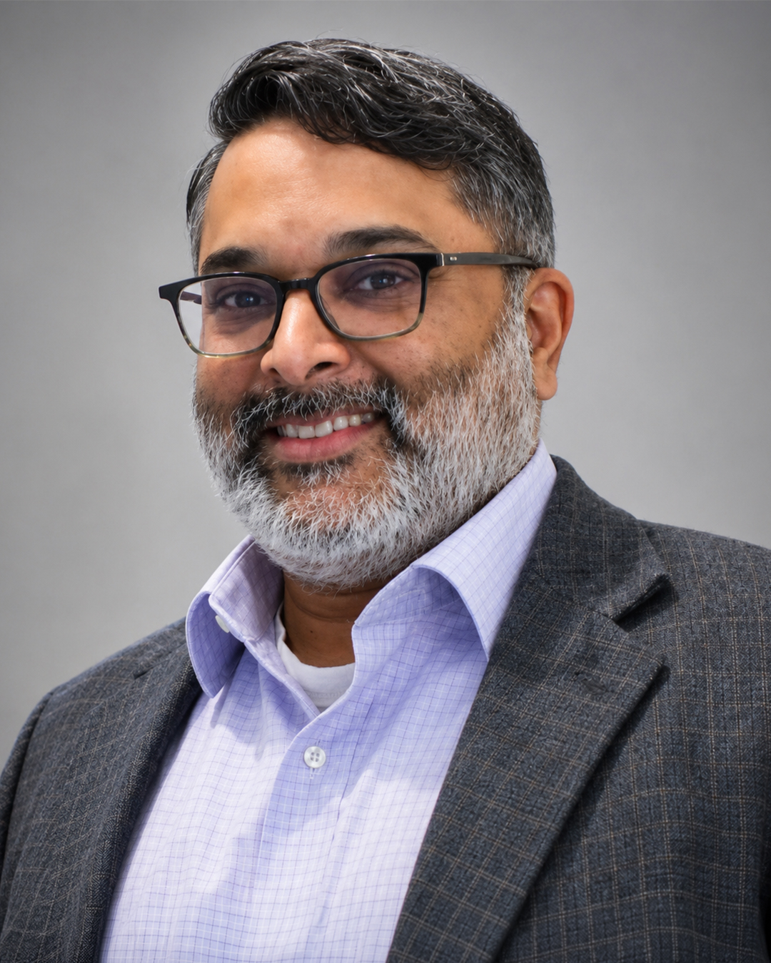Headshot of a smiling man with short dark hair and a salt-and-pepper beard, wearing glasses, a light purple shirt, and a gray blazer, against a neutral background.
