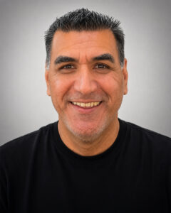 Headshot of a smiling man with short dark hair and light stubble, wearing a black shirt, against a plain gray background.