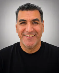 Headshot of a smiling man with short dark hair and light stubble, wearing a black shirt, against a plain gray background.