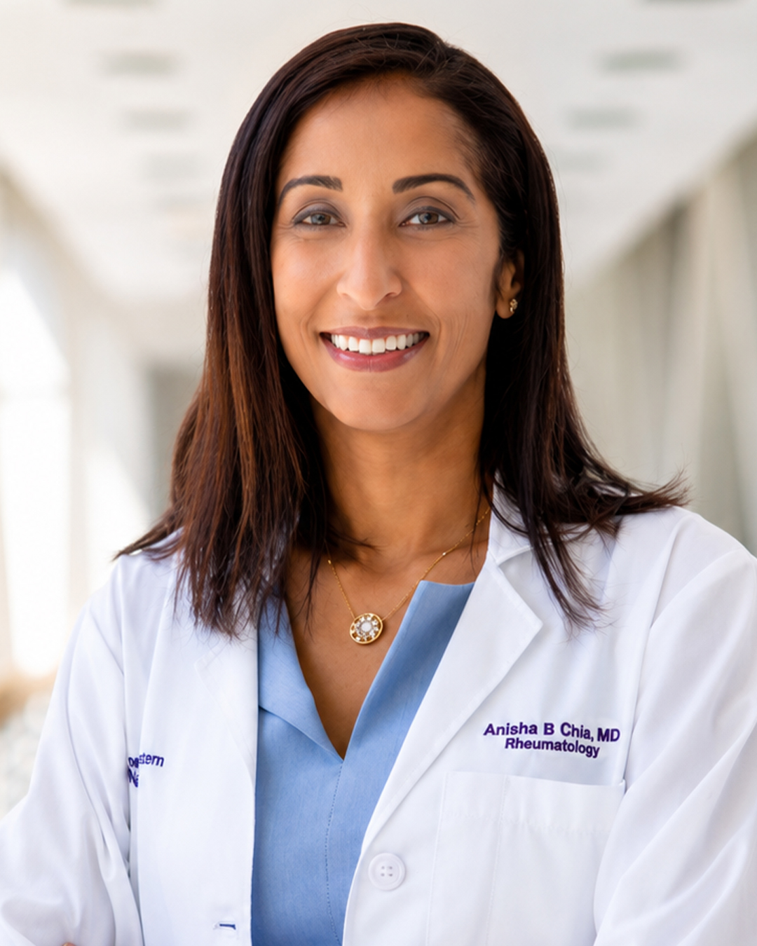 Headshot of a smiling woman wearing a white medical coat labeled “Anisha B. Chia, MD Rheumatology,” over a light blue top, in a clinical setting.