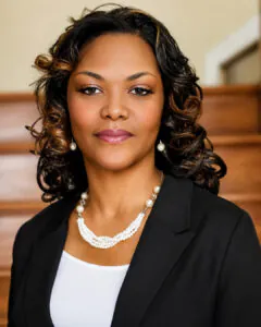 Professional headshot of a woman with shoulder-length curled dark hair wearing a black blazer, white top, and pearl necklace and earrings, standing in front of a softly blurred indoor staircase background.