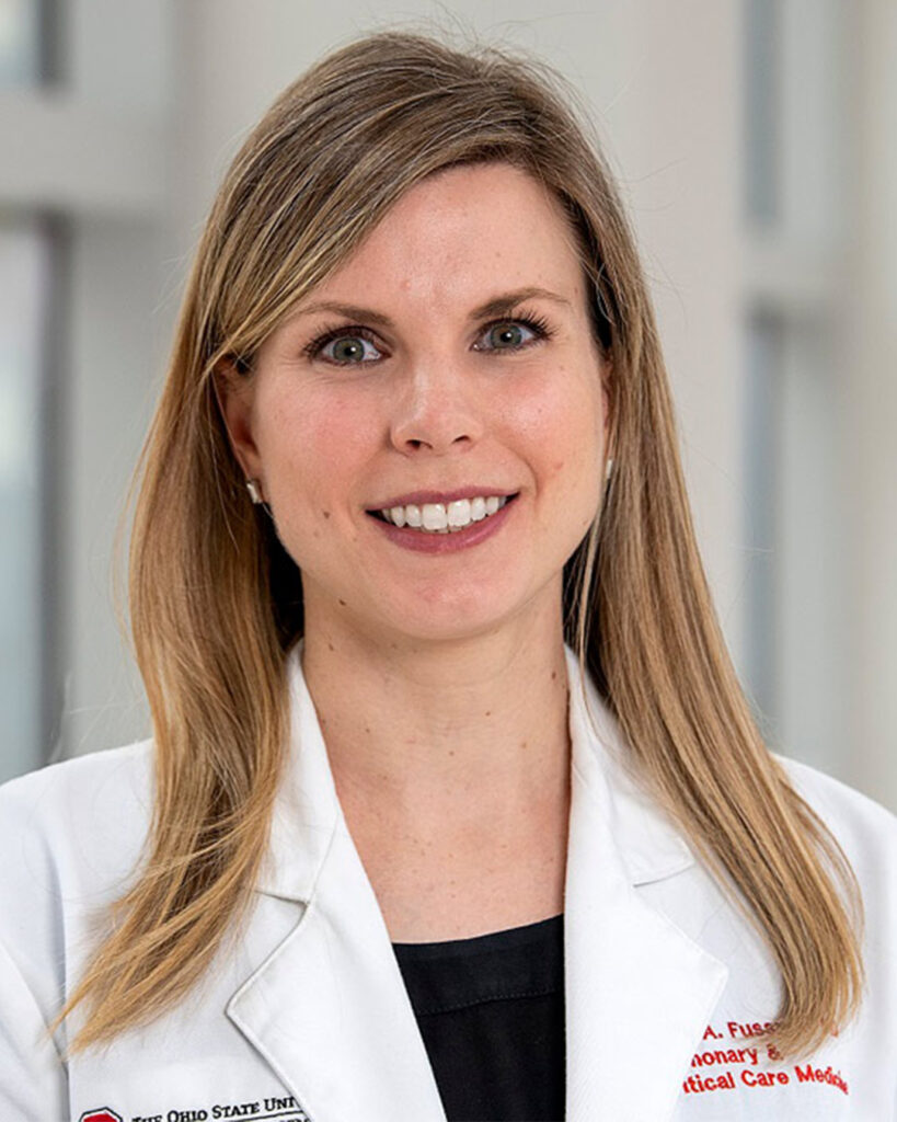 Headshot of a smiling woman with straight blonde hair, wearing a white medical coat, against a softly blurred clinical background.