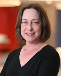 Headshot of a smiling woman with short brown hair, wearing a black top, against a warm-toned blurred indoor background.
