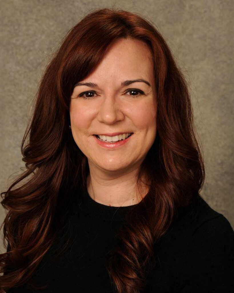 Professional headshot of a smiling woman with long auburn hair wearing a black top, posed against a neutral gray studio background.