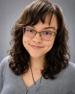 Professional headshot of a smiling young woman with curly dark brown hair and glasses wearing a gray knit sweater and necklace, posed against a soft gray background.