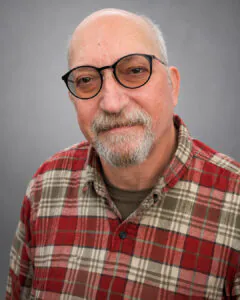 Headshot of an older man with glasses and a gray beard, wearing a red plaid shirt, against a neutral gray background.