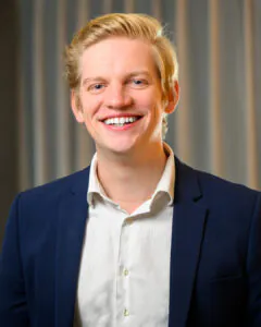 Headshot of a smiling young man with short blonde hair, wearing a white shirt and dark blazer, against a softly blurred striped background.