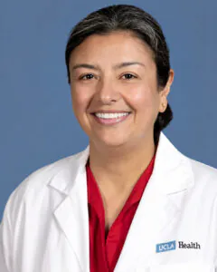Headshot of a smiling woman wearing a white medical coat with a UCLA Health logo over a red blouse, against a blue background.