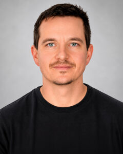 Headshot of a man with short dark hair and light facial hair, wearing a black shirt, against a plain gray background.