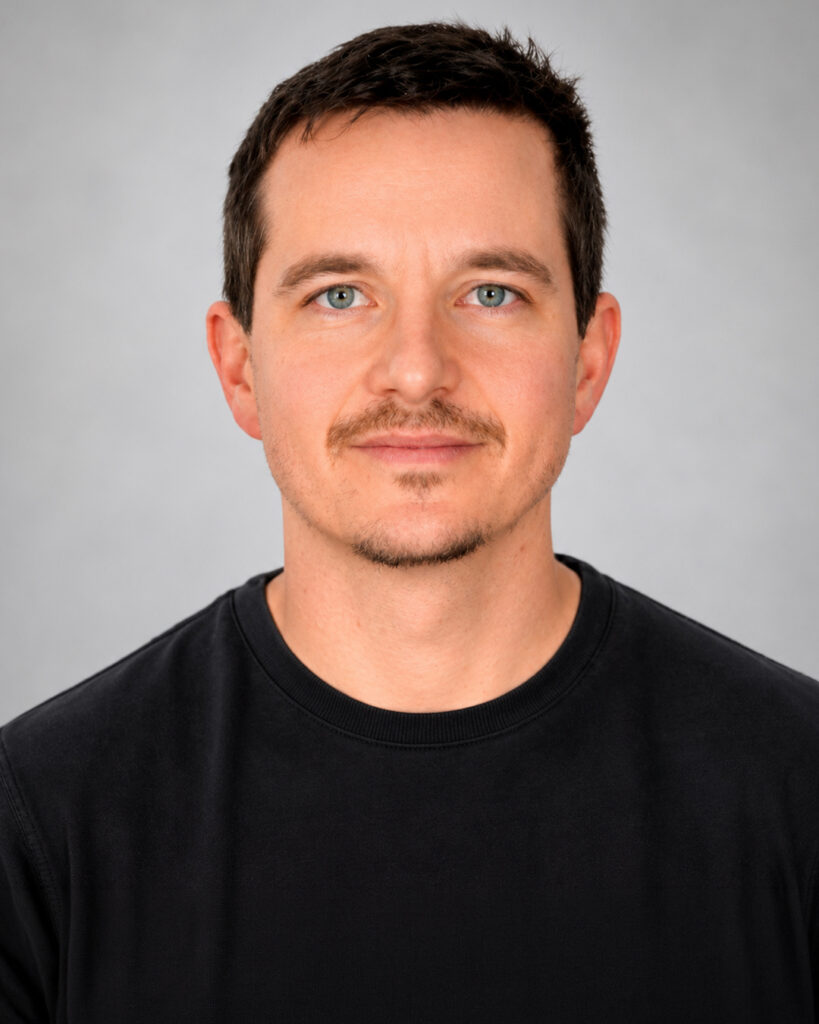 Headshot of a man with short dark hair and light facial hair, wearing a black shirt, against a plain gray background.