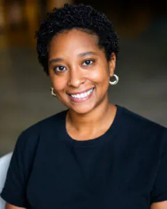 Professional headshot of a smiling Black woman with short curly hair, wearing silver hoop earrings and a black crewneck top. The portrait features soft lighting and a blurred indoor background, creating a polished and approachable appearance.