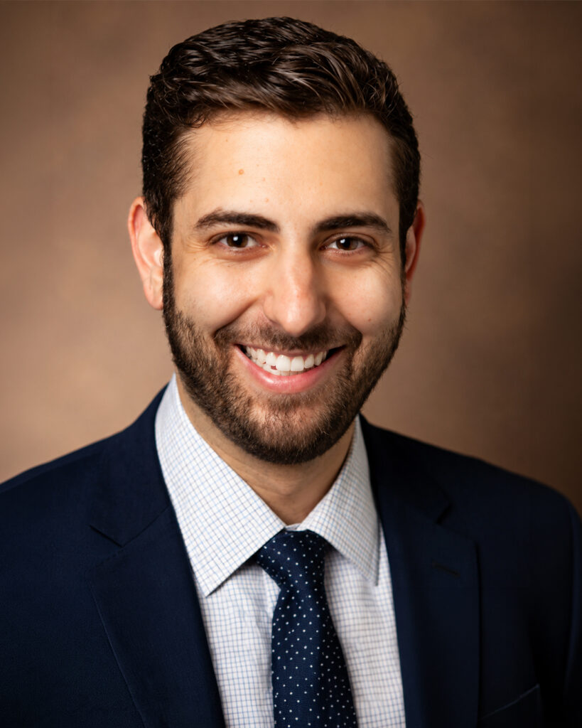 Headshot of a smiling man with dark hair and a beard, wearing a suit and tie, against a warm-toned background.