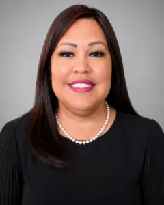 Professional headshot of a smiling woman with straight dark brown hair wearing a black blouse and pearl necklace, posed against a soft gray studio background.