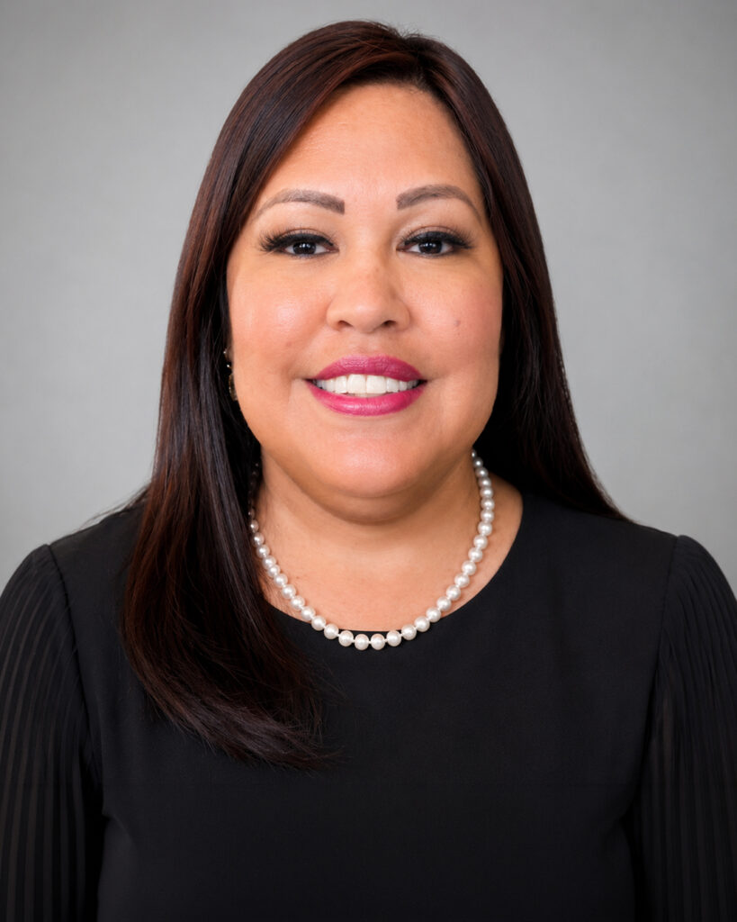Professional headshot of a smiling woman with straight dark brown hair wearing a black blouse and pearl necklace, posed against a soft gray studio background.