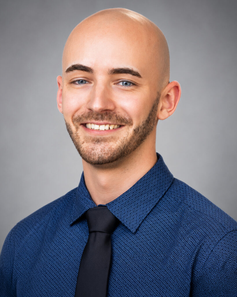 Headshot of a smiling bald man with a short beard, wearing a blue shirt and black tie, against a gray background.