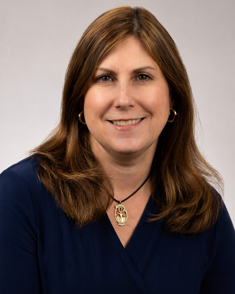 Headshot of a smiling woman with shoulder-length brown hair, wearing a navy top and a pendant necklace, against a neutral background.