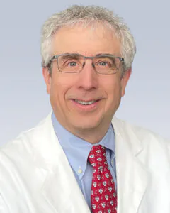 Headshot of a smiling man with gray hair and glasses, wearing a white medical coat and red patterned tie, against a light background.
