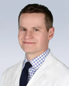 Headshot of a man with short brown hair, wearing a white medical coat, checkered shirt, and dark tie, against a light background.