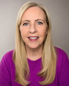 Headshot of a smiling woman with long blonde hair, wearing a bright purple sweater, against a neutral background.