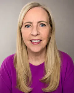 Headshot of a smiling woman with long blonde hair, wearing a bright purple sweater, against a neutral background.