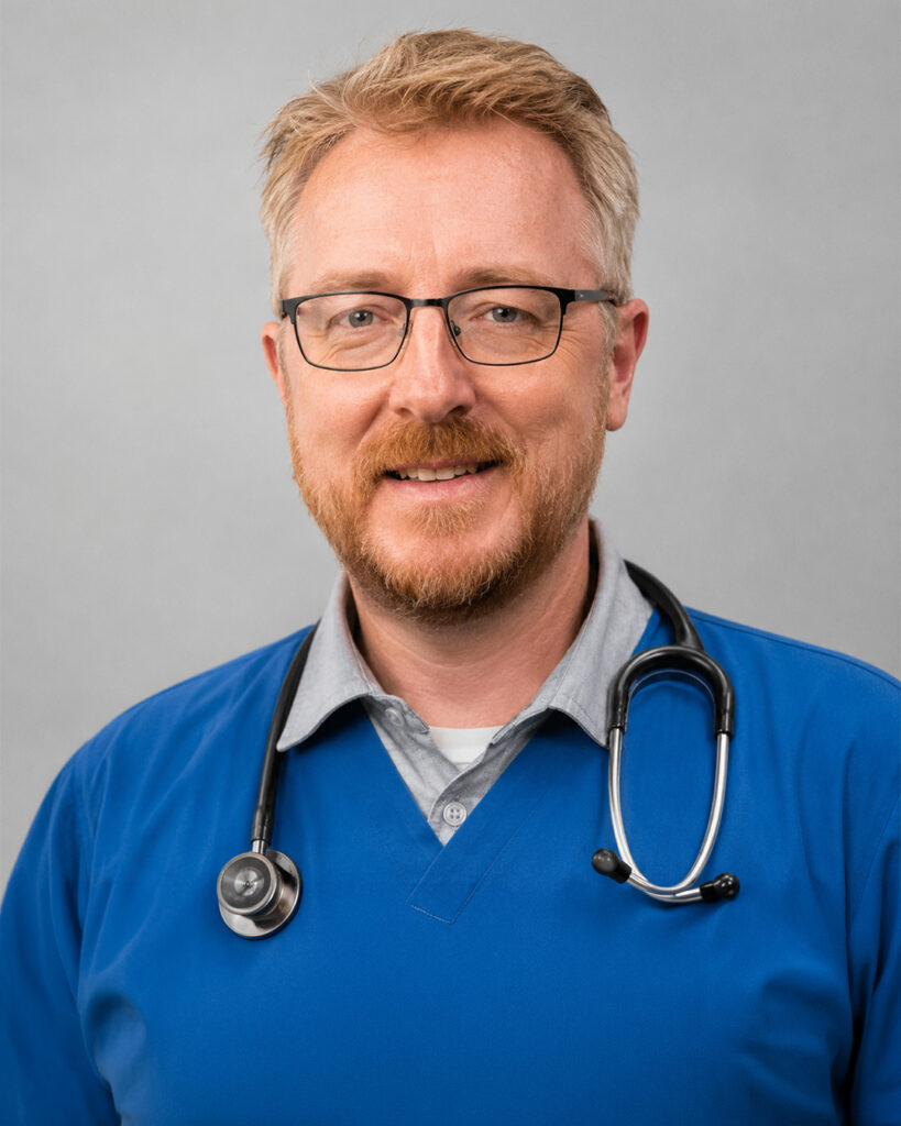 Headshot of a smiling man with light hair, glasses, and a short beard, wearing blue scrubs with a stethoscope, against a neutral background.