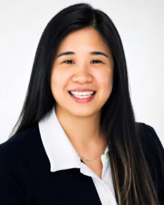 Professional headshot of a smiling Asian woman with long dark hair, wearing a dark blazer over a white collared blouse, against a clean light gray background.
