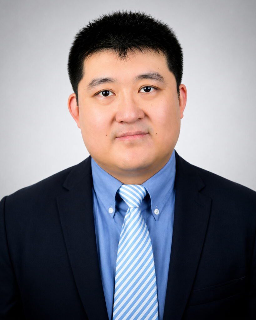 Headshot of a man with short dark hair, wearing a navy suit, blue shirt, and striped tie, against a light gray background.