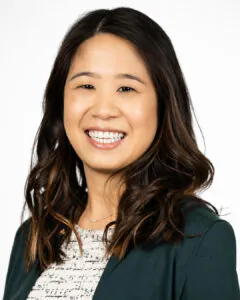 Headshot of a smiling woman with long dark hair, wearing a green blazer over a patterned top, against a light background.