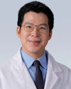 Headshot of a man with short dark hair and glasses, wearing a white medical coat and tie, against a neutral background.