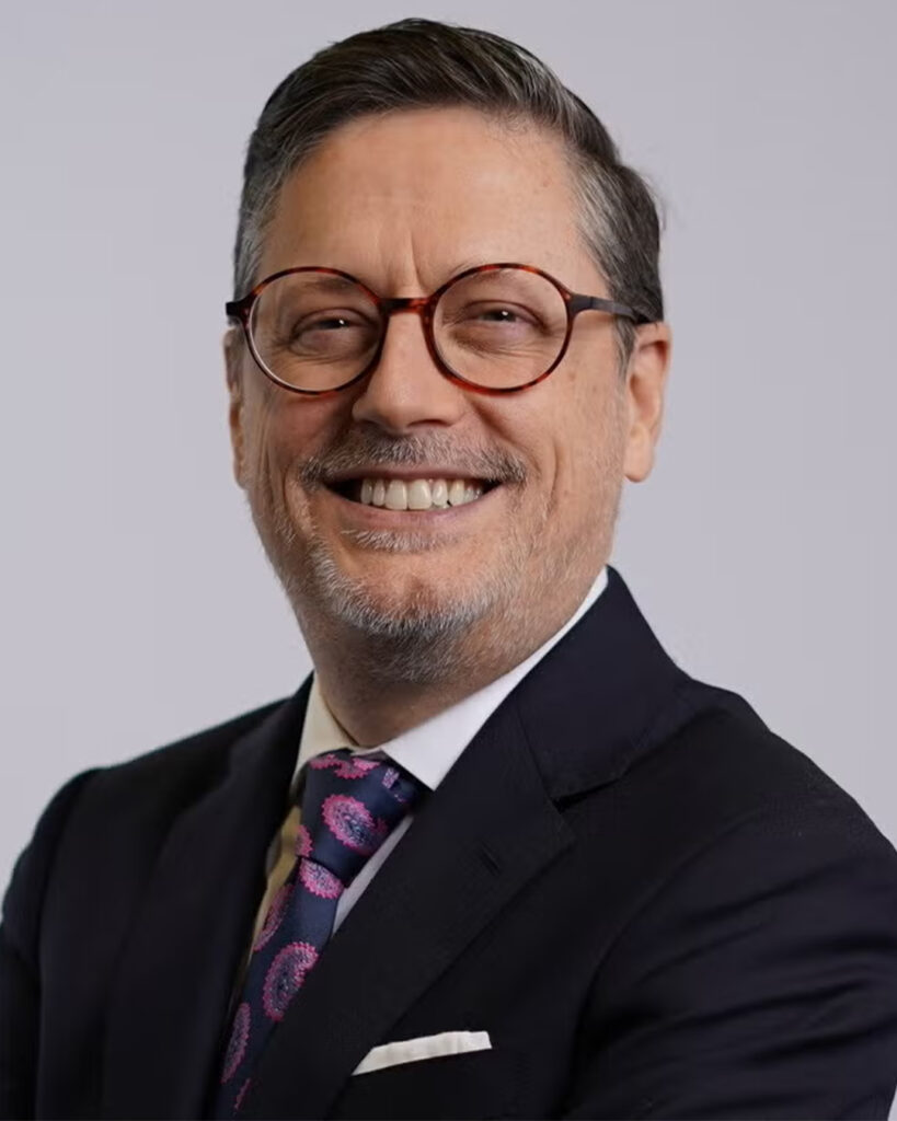 Headshot of a smiling man with gray hair, beard, and round glasses, wearing a dark suit and patterned tie, against a neutral background.