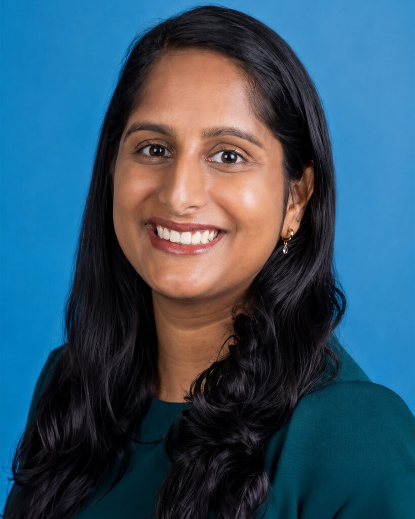 Headshot of a smiling woman with long dark hair, wearing a green top and small earrings, against a blue background.