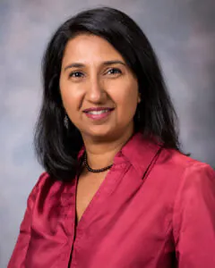 Headshot of a woman with long dark hair, wearing a red blouse and necklace, against a gray background.