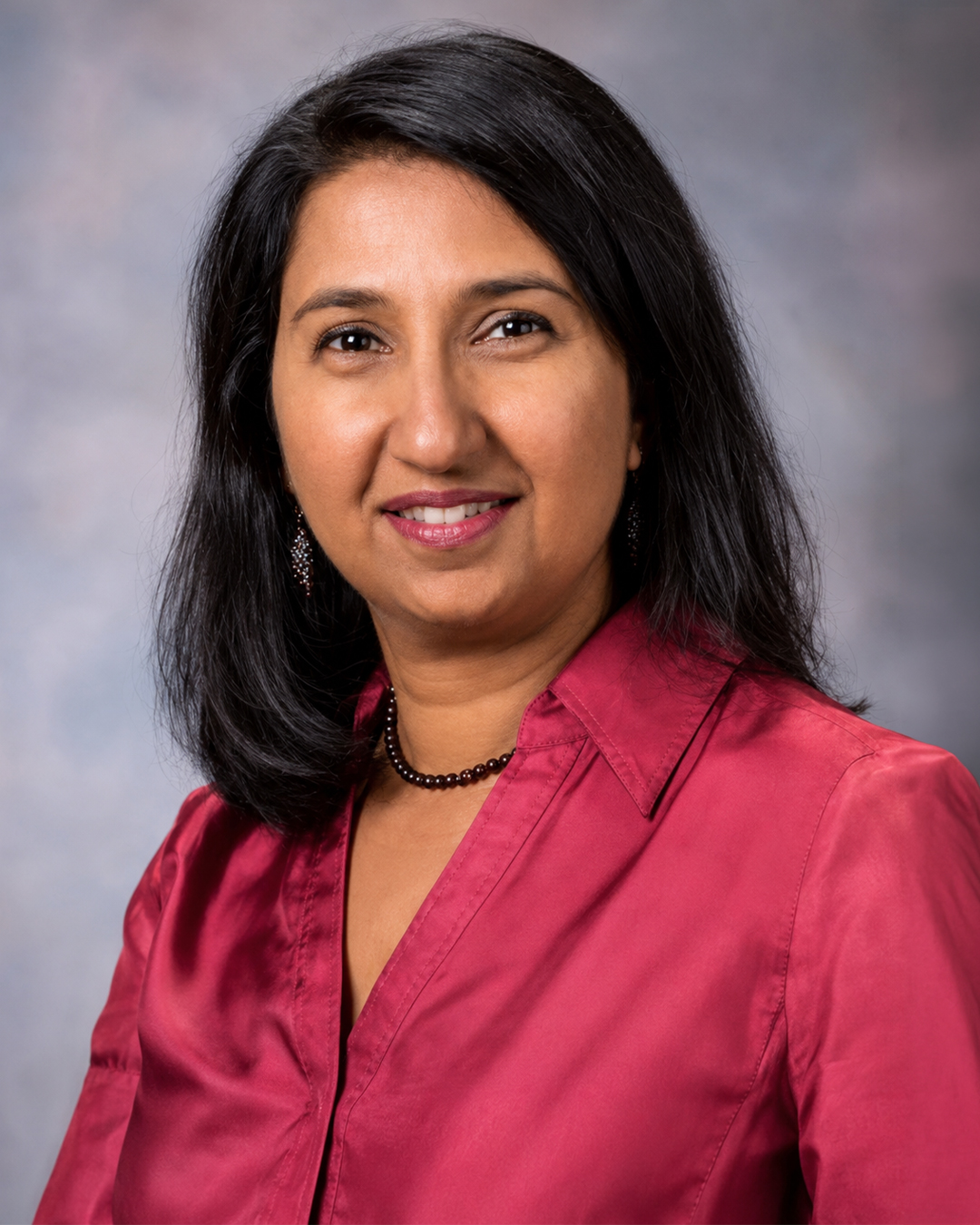 Headshot of a woman with long dark hair, wearing a red blouse and necklace, against a gray background.