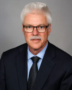Headshot of a man with short white hair, mustache, and glasses, wearing a dark suit and tie, against a neutral background.