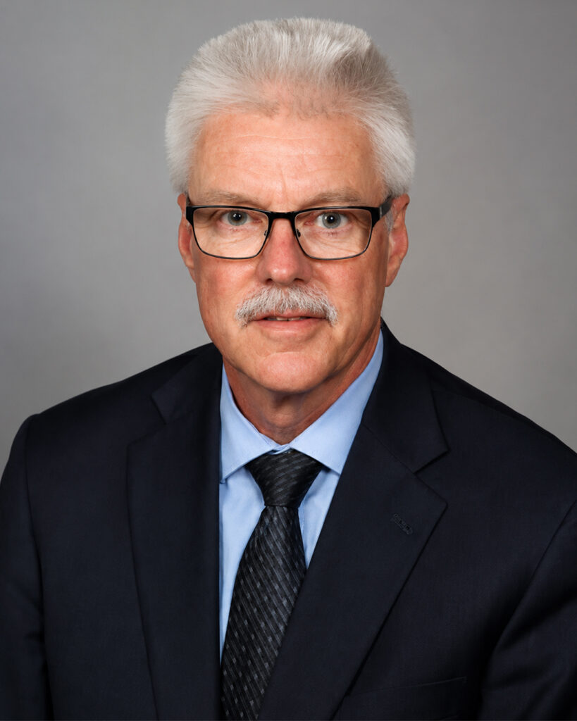 Headshot of a man with short white hair, mustache, and glasses, wearing a dark suit and tie, against a neutral background.