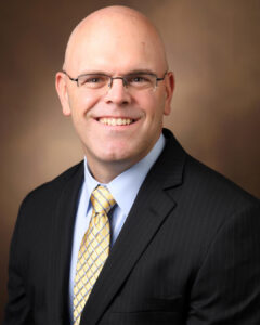 Headshot of a smiling bald man with glasses, wearing a suit and yellow tie, against a warm-toned background.