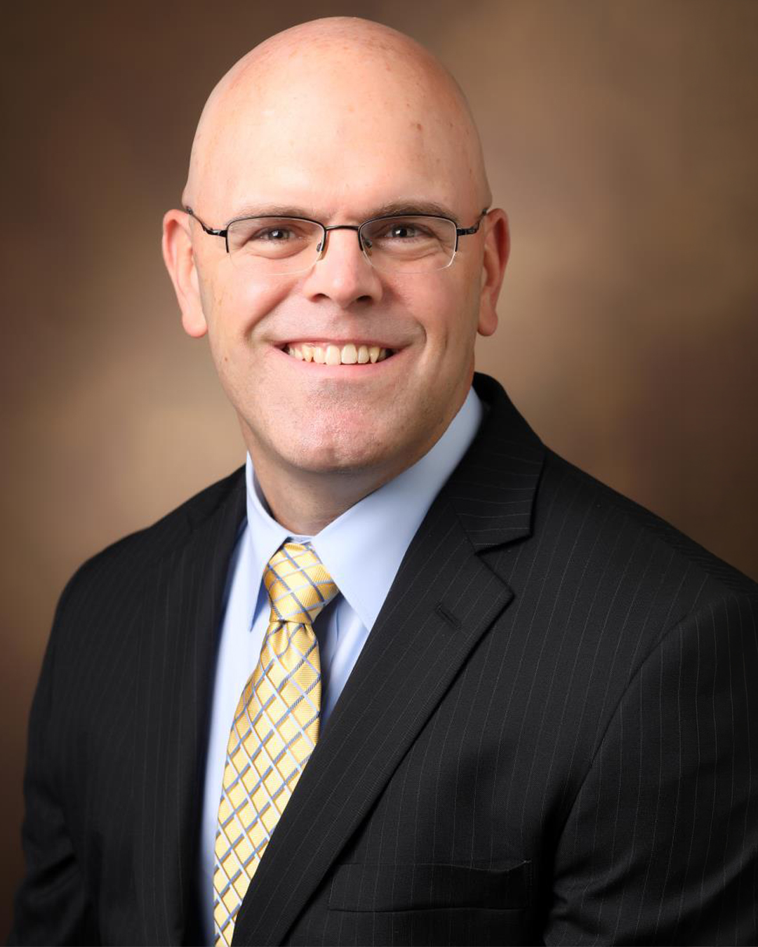 Headshot of a smiling bald man with glasses, wearing a suit and yellow tie, against a warm-toned background.