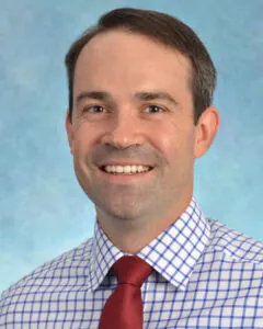 Headshot of a smiling man with short dark hair, wearing a checkered shirt and red tie, against a blue background.
