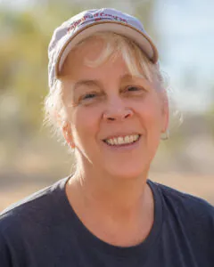 Professional outdoor headshot of an older woman smiling at the camera while wearing a light-colored baseball cap and dark gray T-shirt. The background is softly blurred with trees and warm natural lighting, creating a relaxed and approachable portrait.