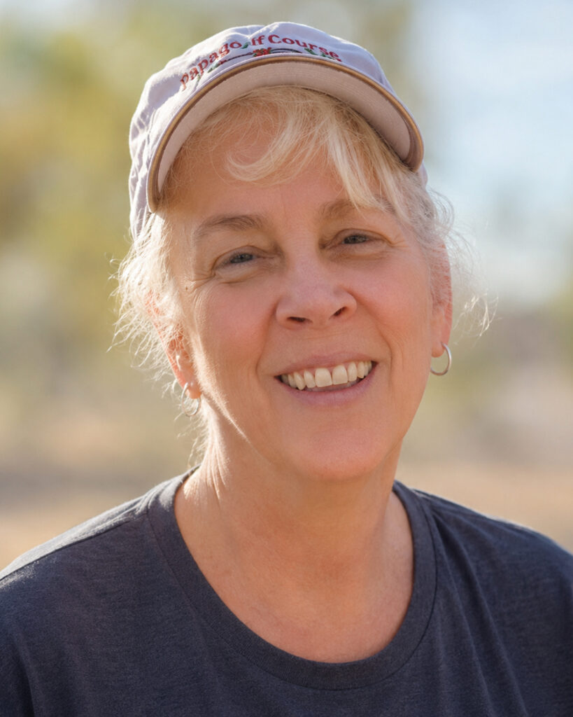 Professional outdoor headshot of an older woman smiling at the camera while wearing a light-colored baseball cap and dark gray T-shirt. The background is softly blurred with trees and warm natural lighting, creating a relaxed and approachable portrait.